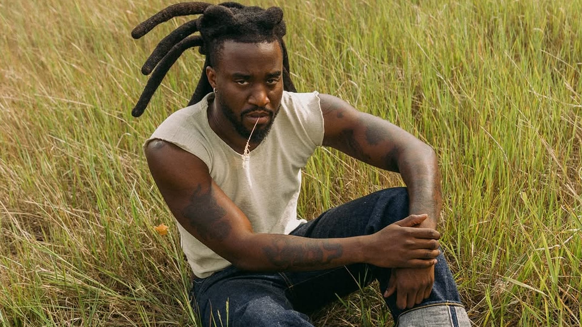 Shaboozey sits in tall Montana grass wearing a sleeveless shirt and jeans, facing backlash after a photo shoot allegedly took place on private land without permission.