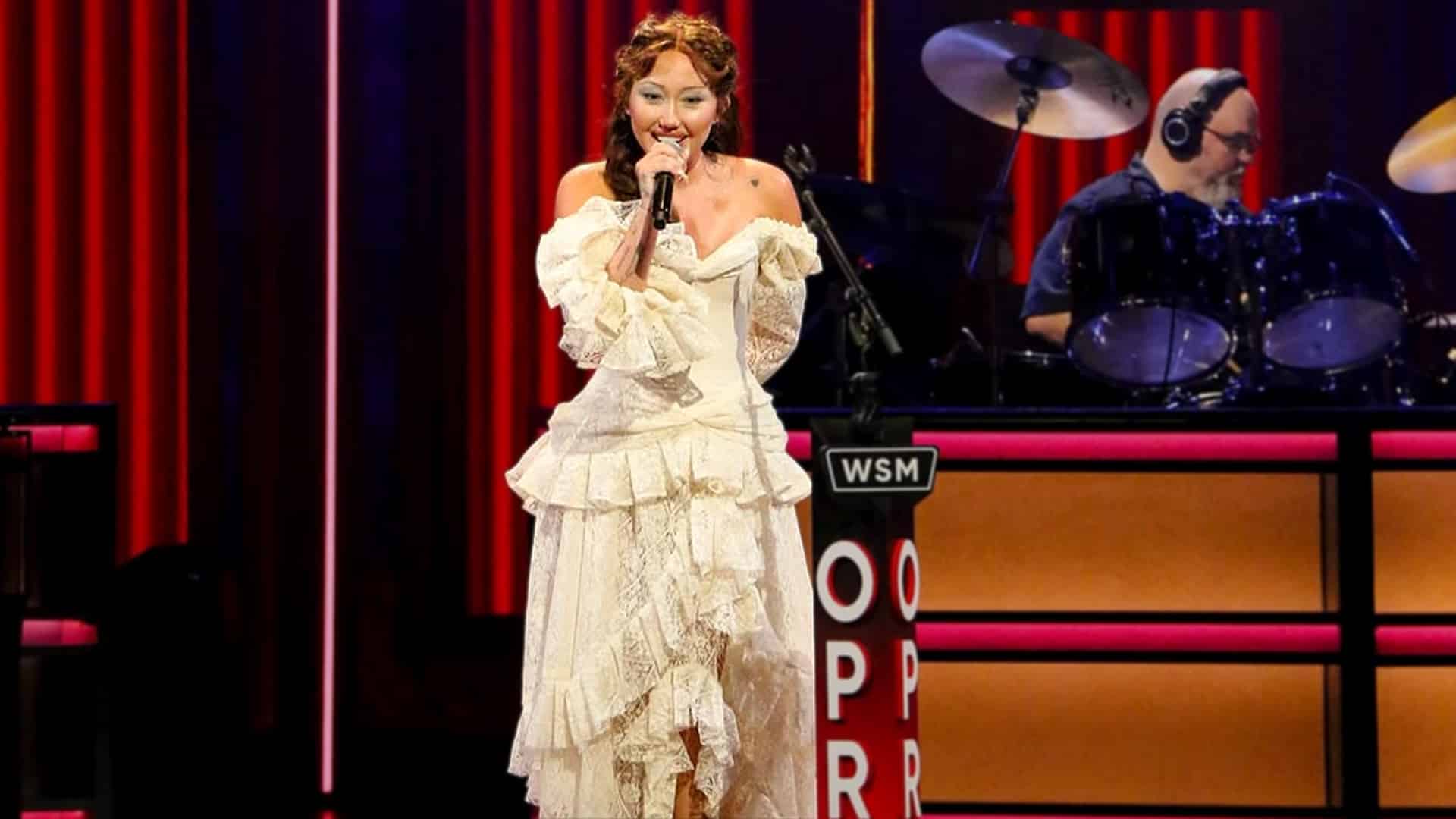 Noah Cyrus stands on the Grand Ole Opry stage in a white lace dress, singing into a microphone with a drummer playing behind her.