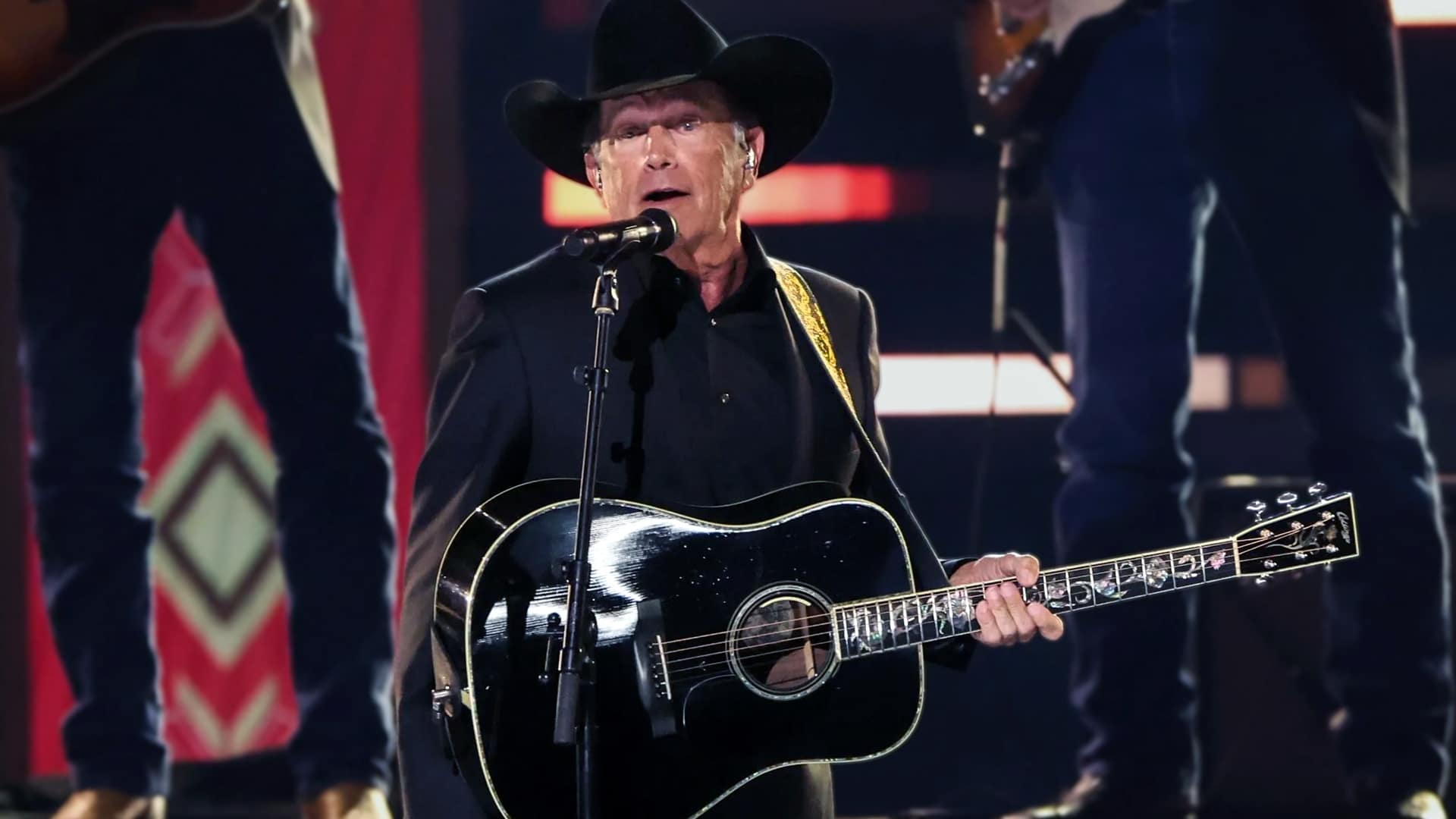 George Strait performs on stage in his signature black cowboy hat and suit, playing a black acoustic guitar. His name was falsely used in a Facebook flood benefit scam.