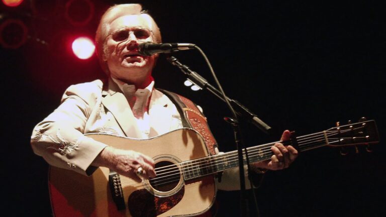 Country legend George Jones plays guitar under spotlight, embodying the raw heartbreak behind these 12 most depressing country songs.