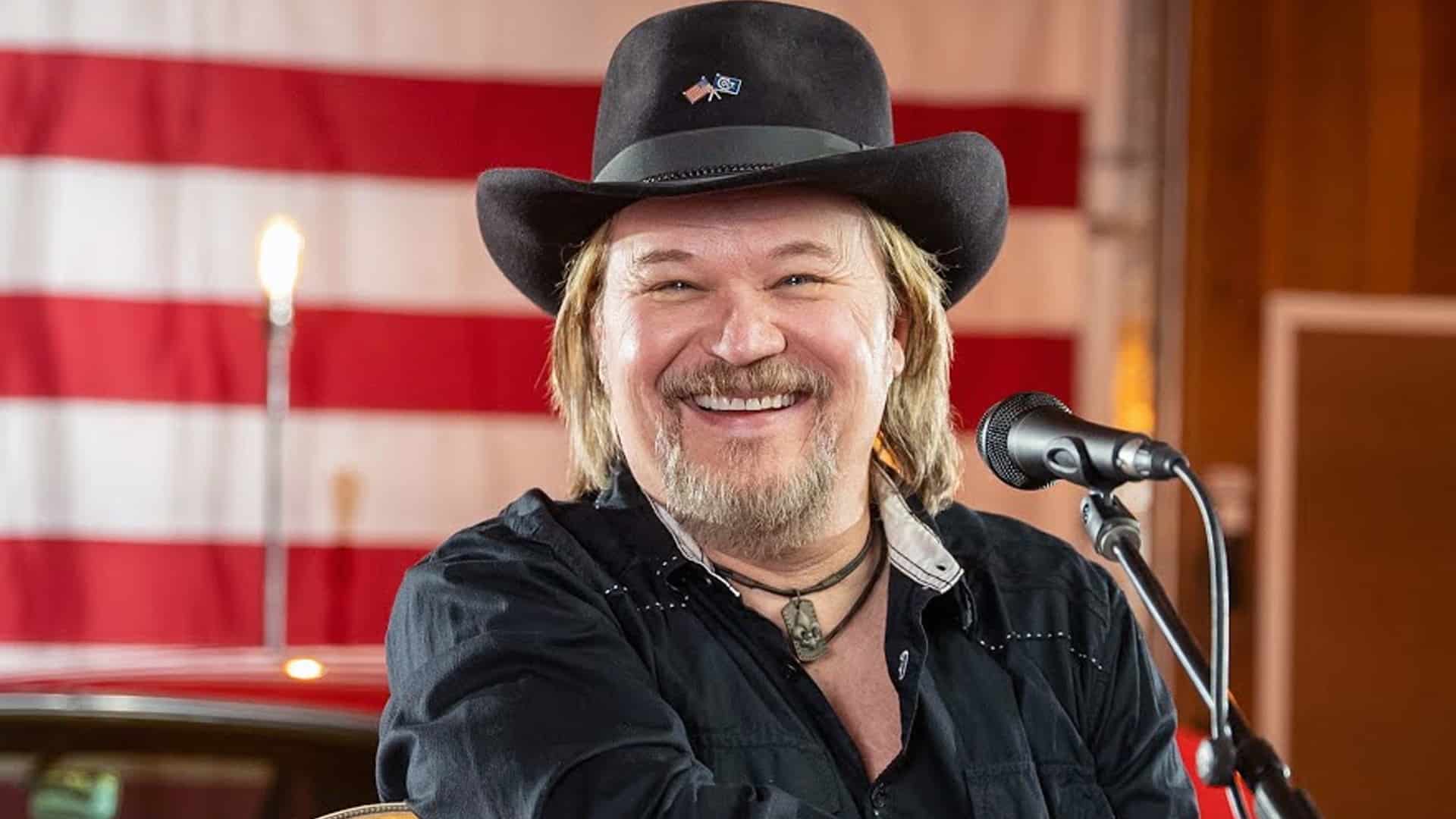 Travis Tritt smiles in a black cowboy hat and shirt while performing "Good Ol' Boys" with a microphone in front of an American flag backdrop.