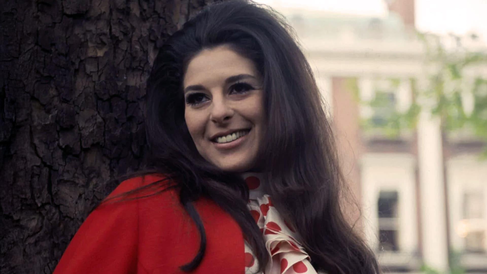 Bobbie Gentry smiles in a red coat beside a tree, capturing the mystique of the country star who vanished from the spotlight.