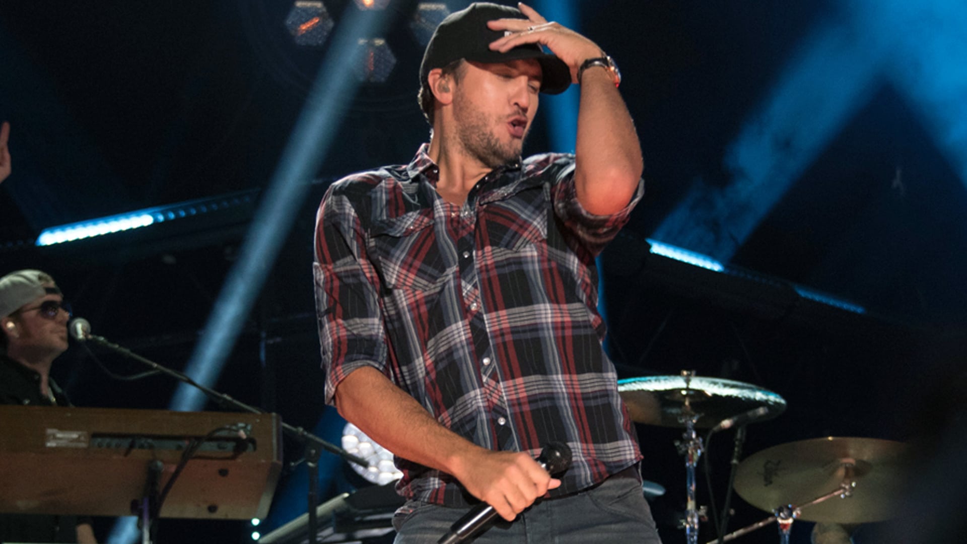 Luke Bryan performs with high energy on stage in a plaid shirt and cap, dividing fans over whether he's saving or ruining country music.