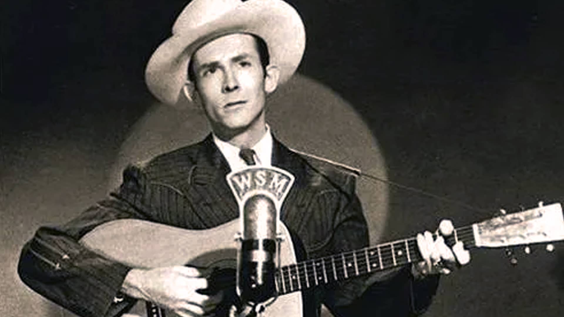 Hank Williams performs in a cowboy hat and suit, playing guitar before a WSM microphone during his early Grand Ole Opry days.