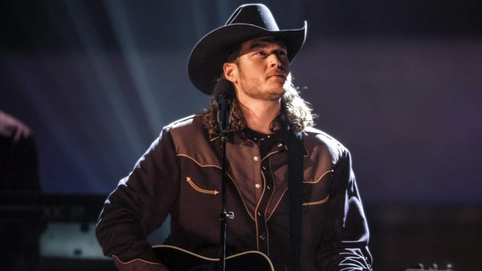 A young Blake Shelton in a cowboy hat plays guitar on stage, recalling how songwriter Mae Boren Axton inspired him to move to Nashville at 17.