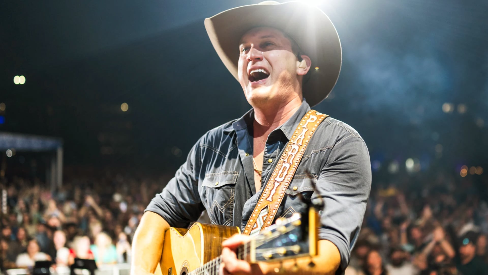 Jon Pardi performs on stage with a guitar, wearing a cowboy hat and smiling under bright lights, ahead of his 2025 Big 12 Championship halftime show.