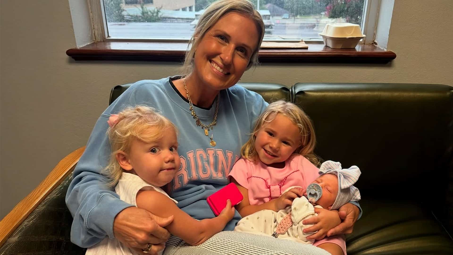 Korie Robertson beams with joy as she sits on a couch holding granddaughters Honey, Haven, and newborn Kit Carroway Huff, all smiling and snuggled up together.