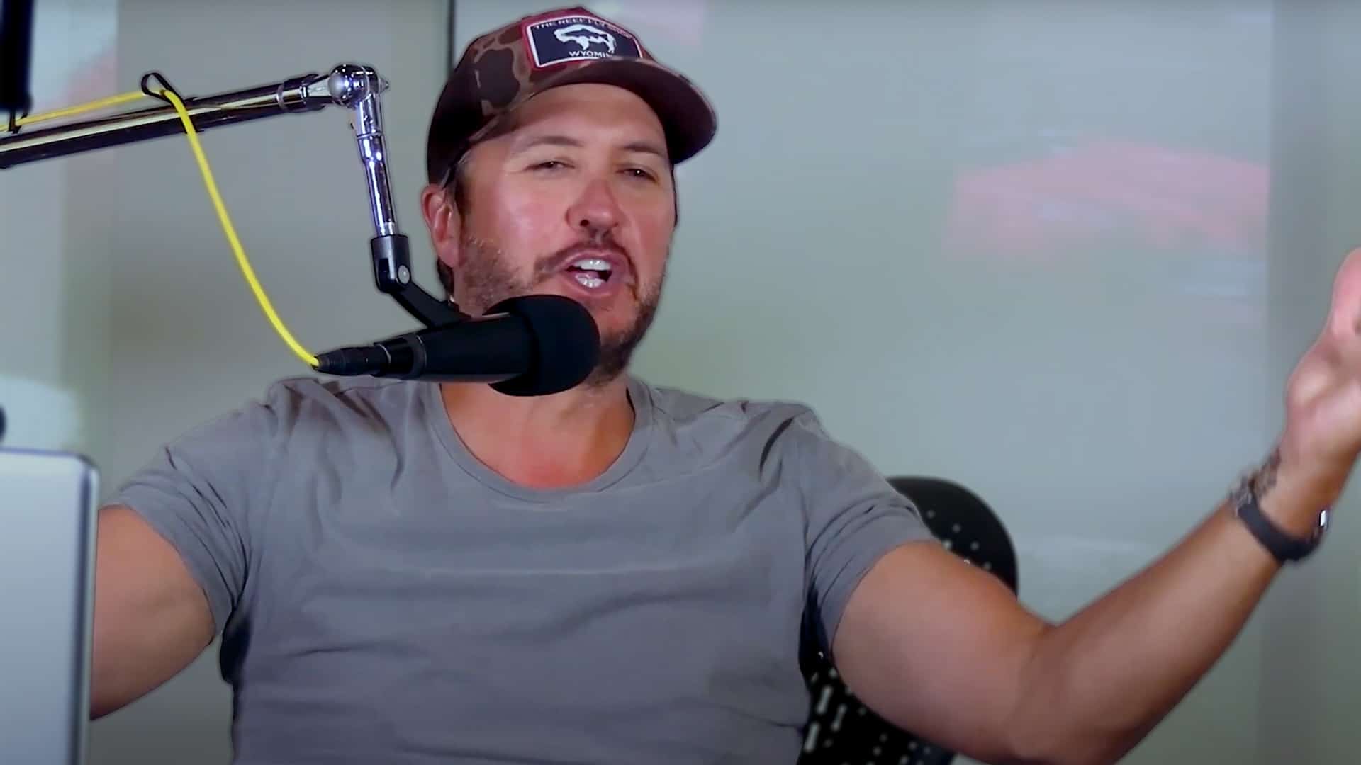 Luke Bryan speaking energetically into a podcast microphone, wearing a gray shirt and camo cap, as he talks about being more unfiltered and bold on stage.