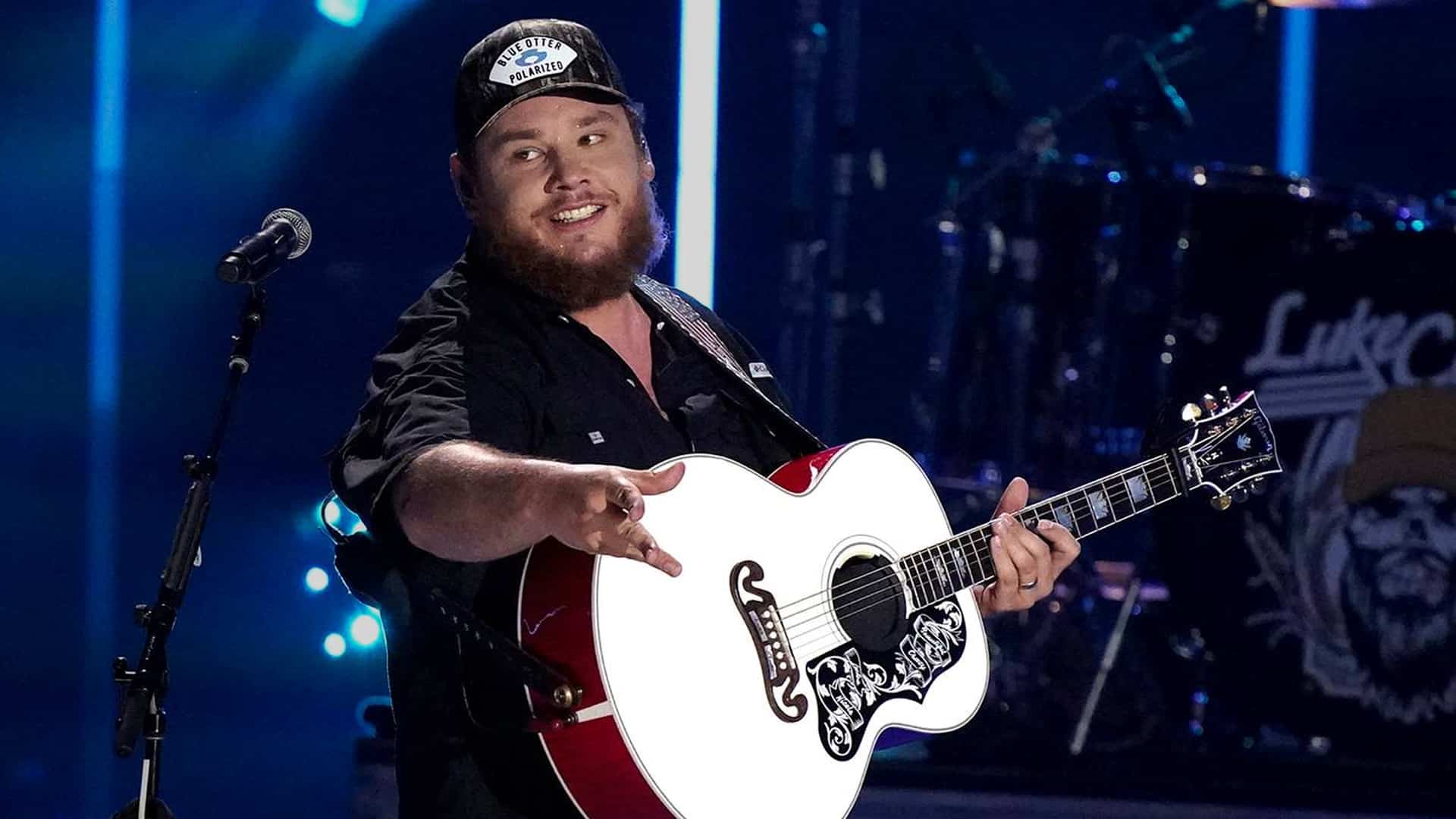Luke Combs smiles on stage mid-performance, wearing a cap and strumming his signature white guitar, teasing fans with a bold idea for his next album.
