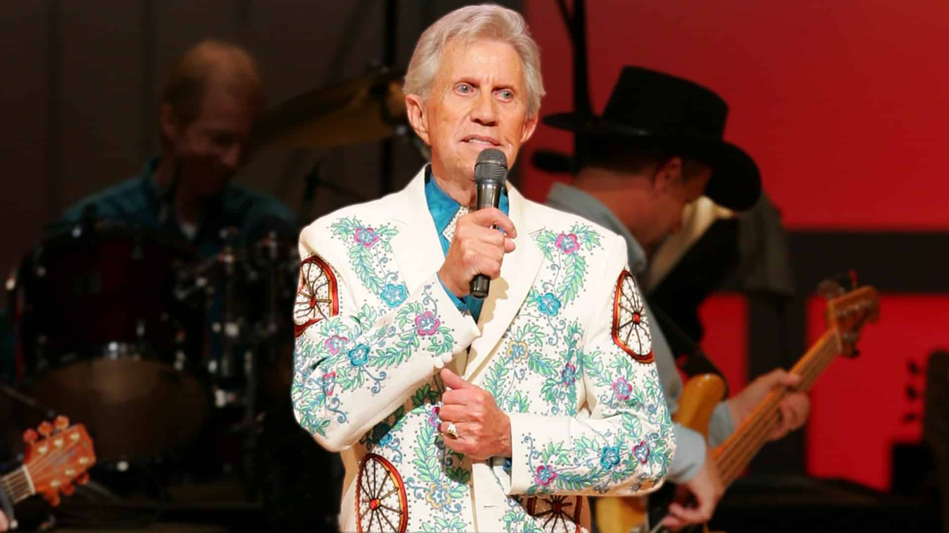 Porter Wagoner holds a microphone on stage, wearing a white rhinestone suit adorned with colorful floral patterns and wagon wheel designs.