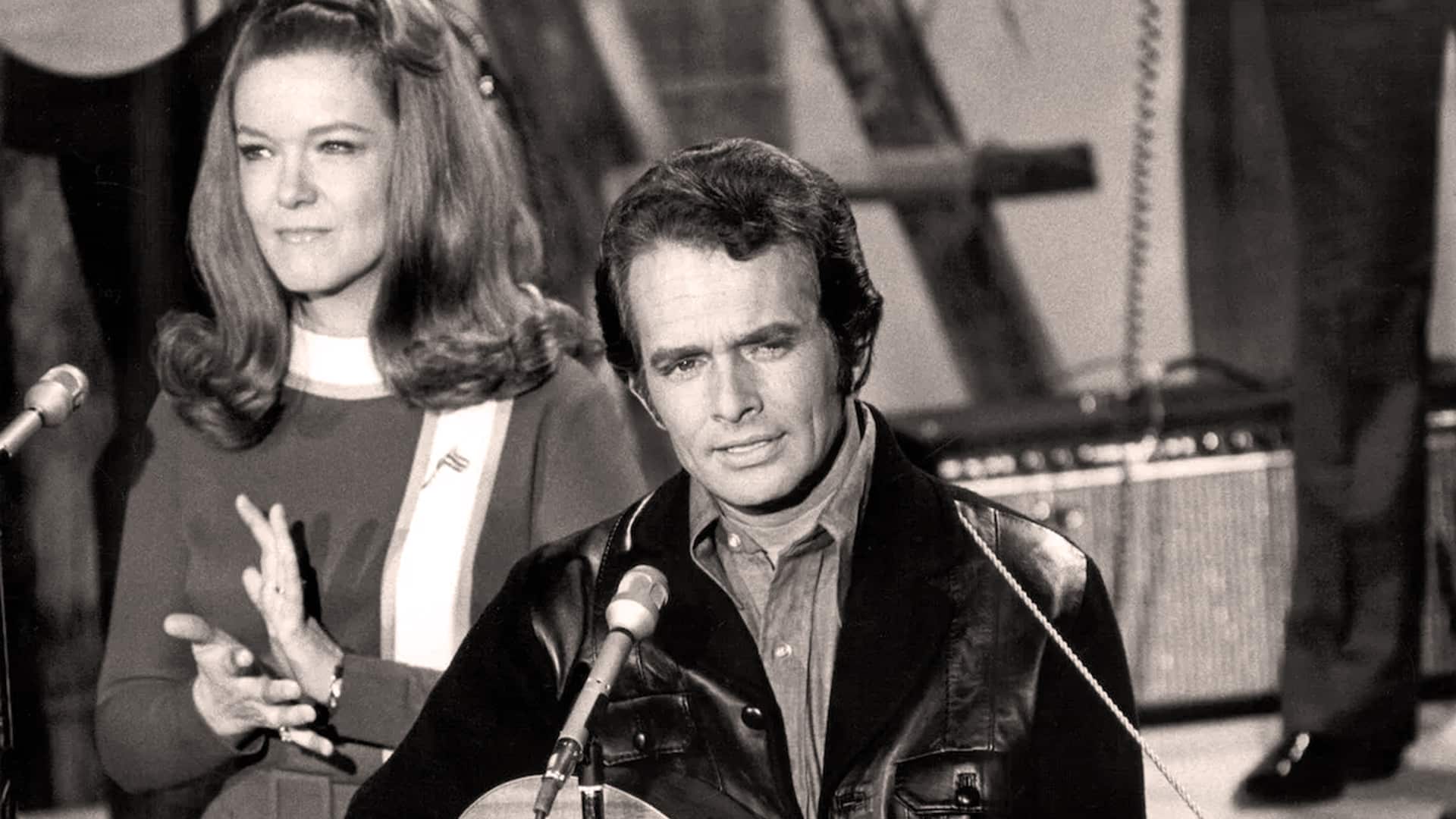 Merle Haggard sings into a microphone with guitar in hand, while Bonnie Owens stands beside him, showing the partnership that helped inspire classics like “Mama Tried” and “Today I Started Loving You Again.”
