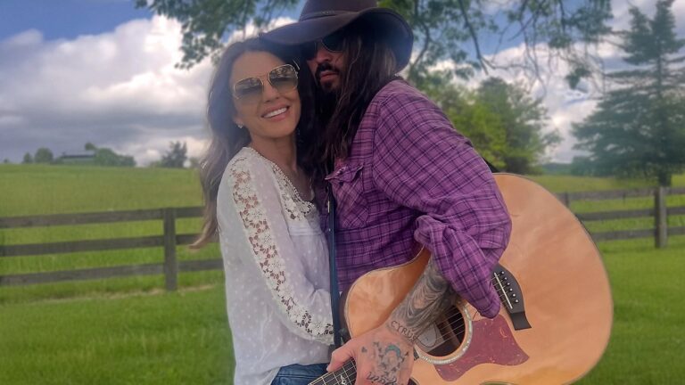 Elizabeth Hurley smiles beside Billy Ray Cyrus holding a guitar, marking their playful summer of music and fresh beginnings.