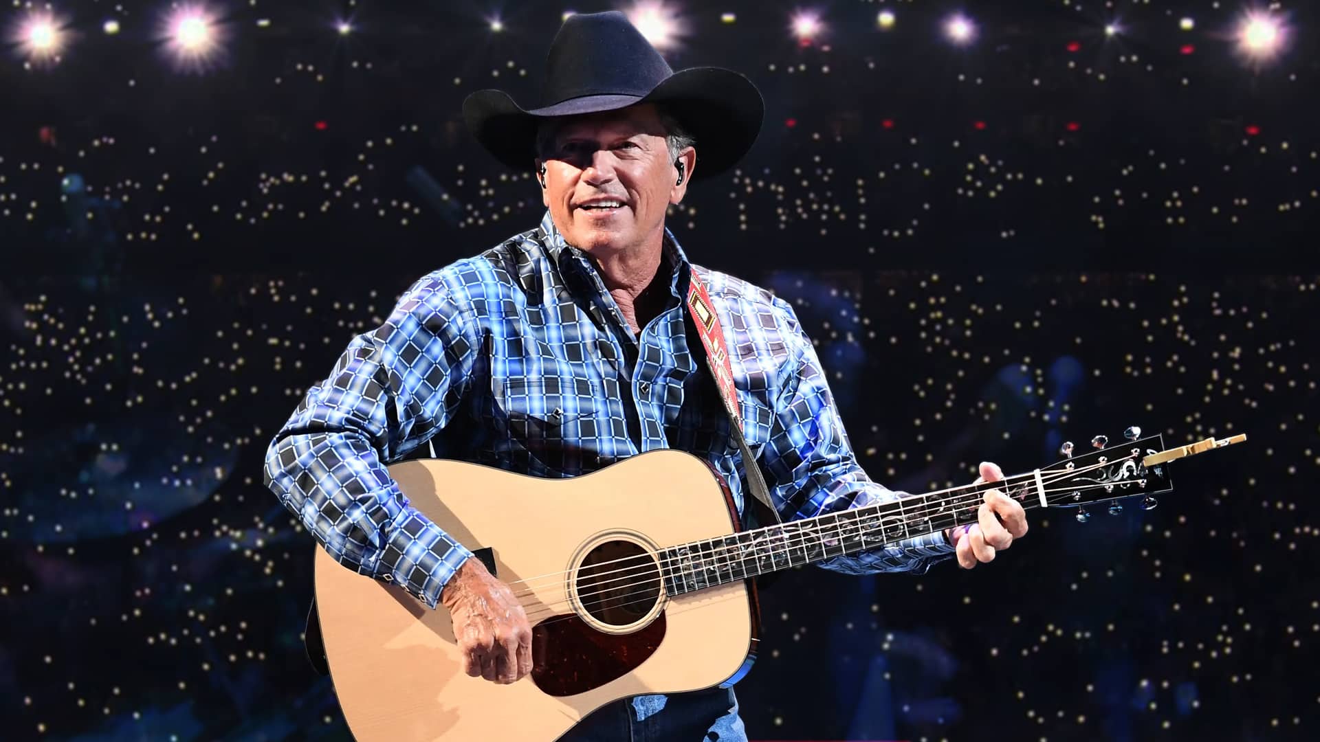 Country legend George Strait on stage in a cowboy hat and plaid shirt, smiling as Super Bowl 2026 rumors swirl around his latest collaboration.