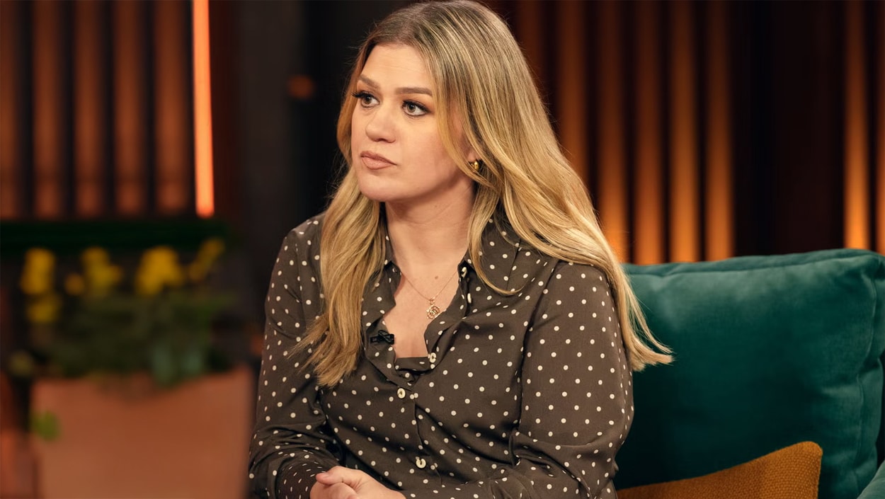 Kelly Clarkson seated in a studio, wearing a polka-dot blouse and appearing reflective while addressing fans online.