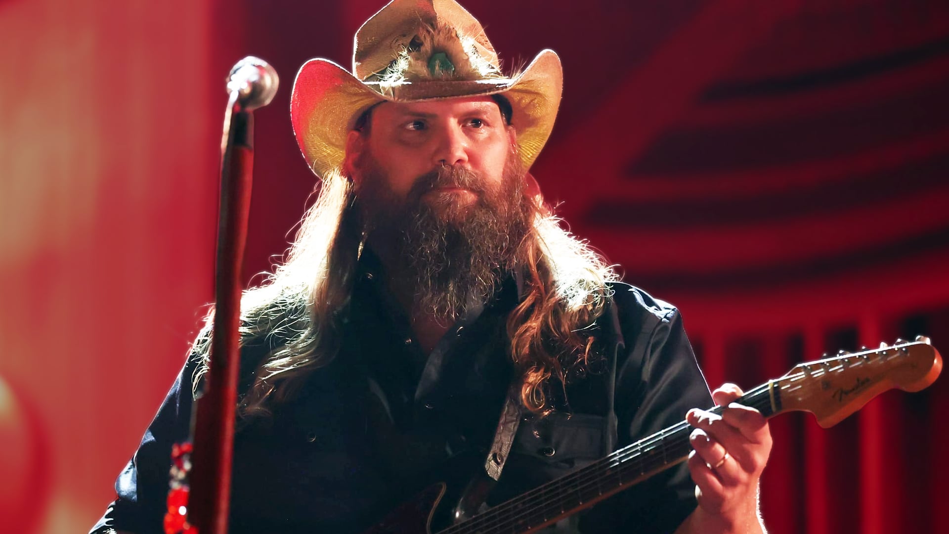 Country singer Chris Stapleton playing guitar under warm stage lights, following the announcement that he’s postponing shows to recover his voice.
