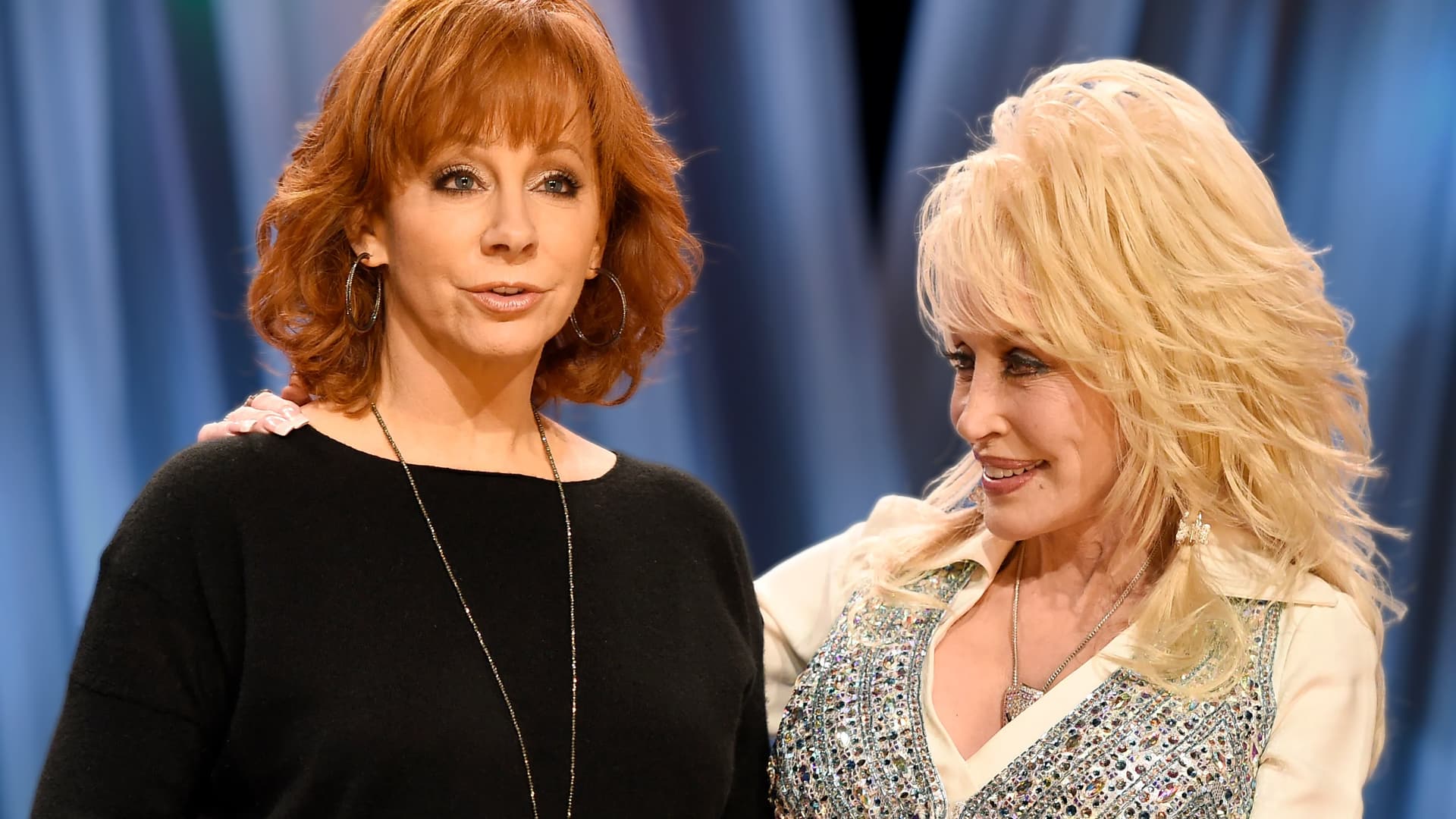 Country icons Reba McEntire and Dolly Parton standing side by side, smiling as Reba delivers a witty response to viral fake photo rumors.