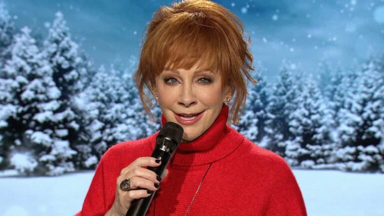 Reba McEntire, smiling with a microphone against a snowy backdrop, is ready to host and perform at this year’s Christmas in Rockefeller Center.