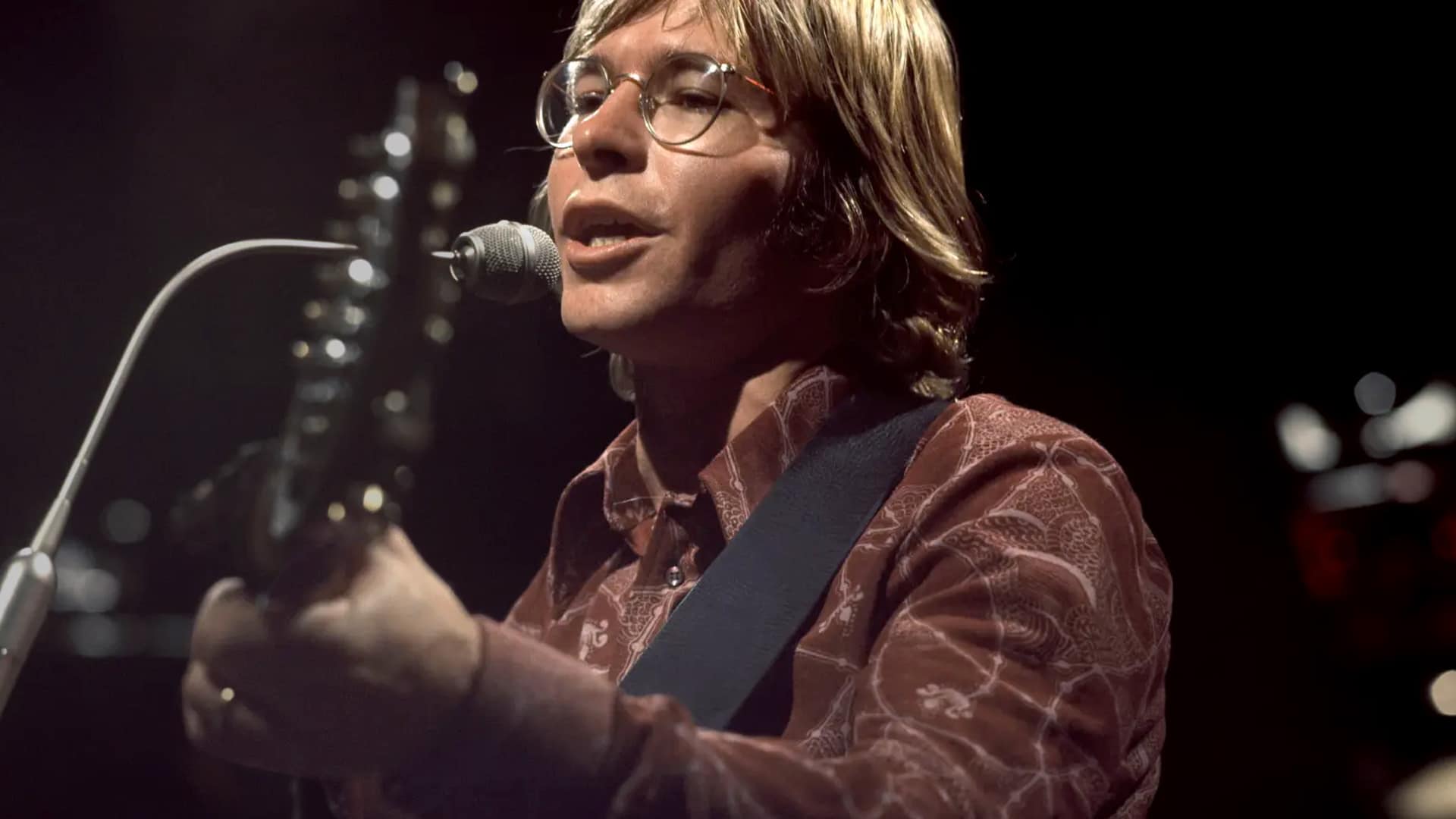 ohn Denver performing with his guitar and signature glasses, a reminder of the soulful voice lost in the 1997 plane crash tragedy.
