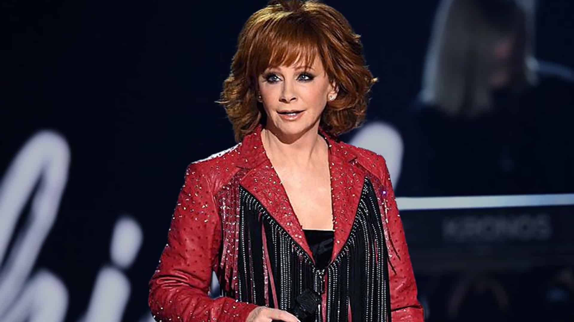 Reba McEntire performs on stage in a red rhinestone jacket, holding a microphone during a concert, a reminder of the heartwarming story from 30 years ago when she stopped a show to honor a special needs fan.