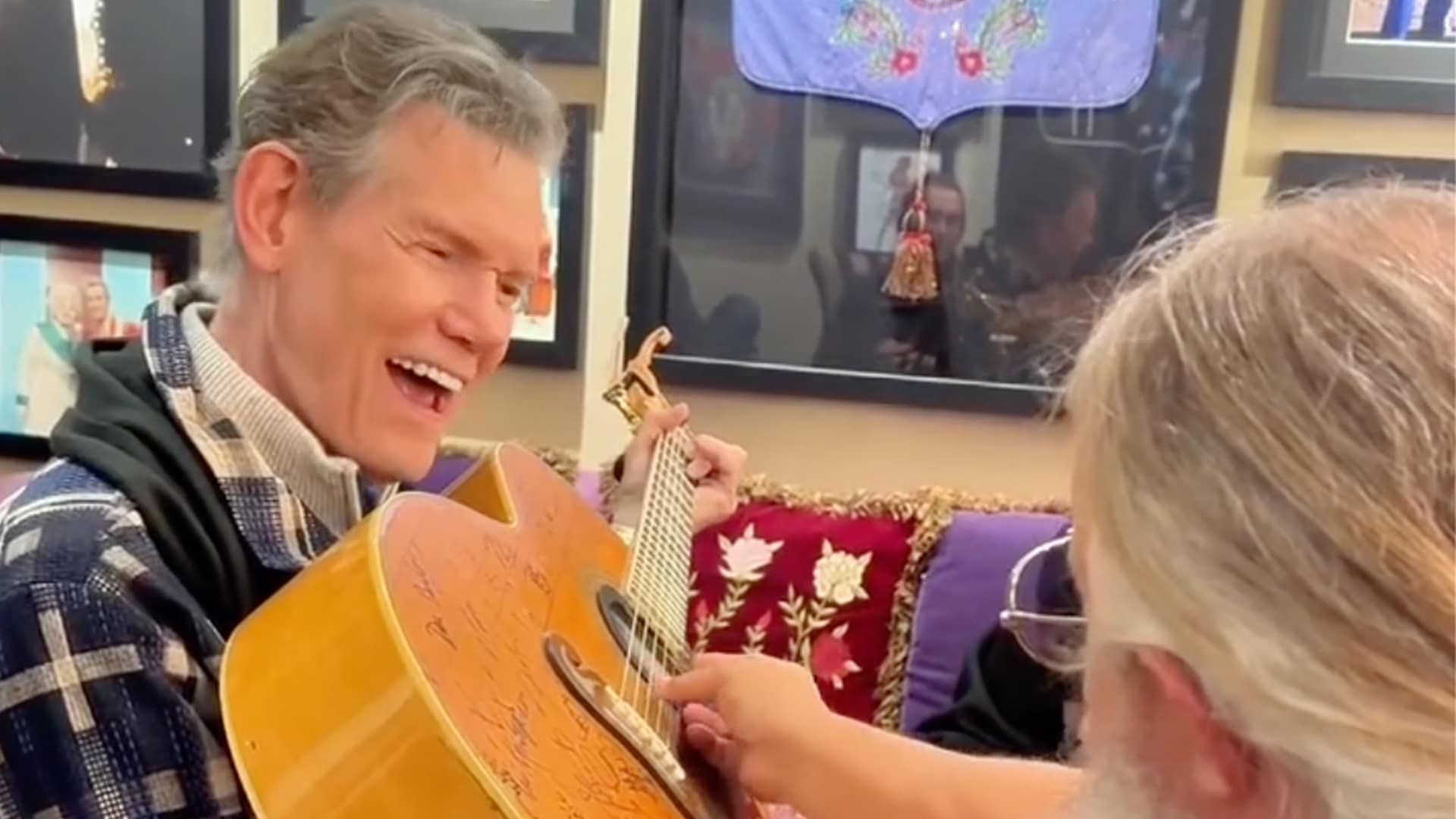 Randy Travis is holding a signed guitar while Jamey Johnson guides his hand across the strings, capturing the tender backstage moment that showed his enduring spirit.