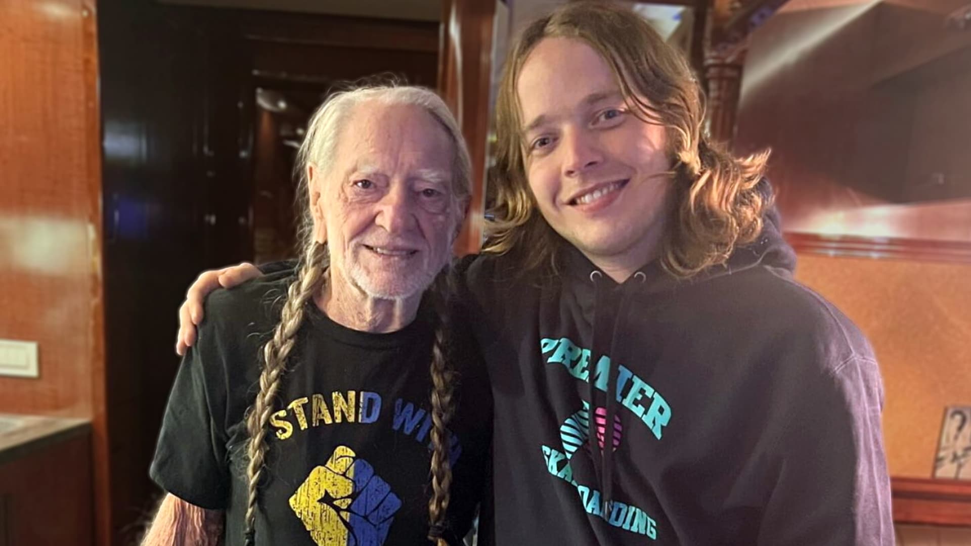 Billy Strings smiling with long hair and Premier Trading hoodie next to Willie Nelson with braid and Stand With fist shirt after Billy lost $1000 poker game and jammed on Trigger priceless night.