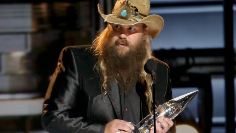 Chris Stapleton with long beard and hair in feathered brown cowboy hat and black suit jacket holding CMA trophy and speaking into microphone during acceptance speech at Country Music Association Awards.