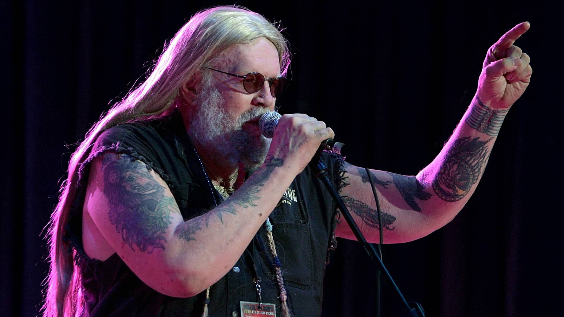 Country singer David Allan Coe sings into a microphone with one arm raised, showing his tattoos during a live performance, reflecting on his complex and controversial legacy.