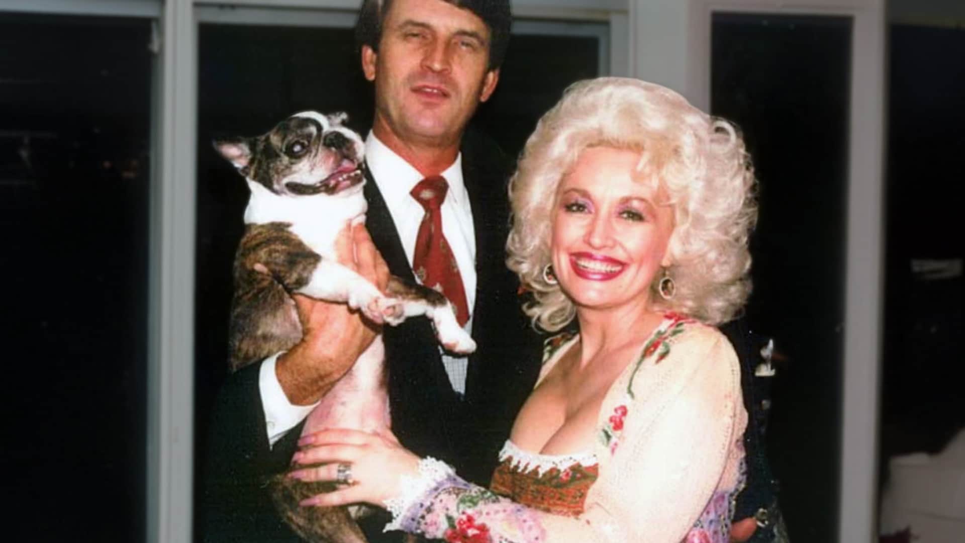 Young Dolly Parton in floral dress with big blonde hair holding small dog next to Carl Dean in suit vintage photo from early marriage showing humble Sears wedding ring Dolly kept forever instead of upgrading.