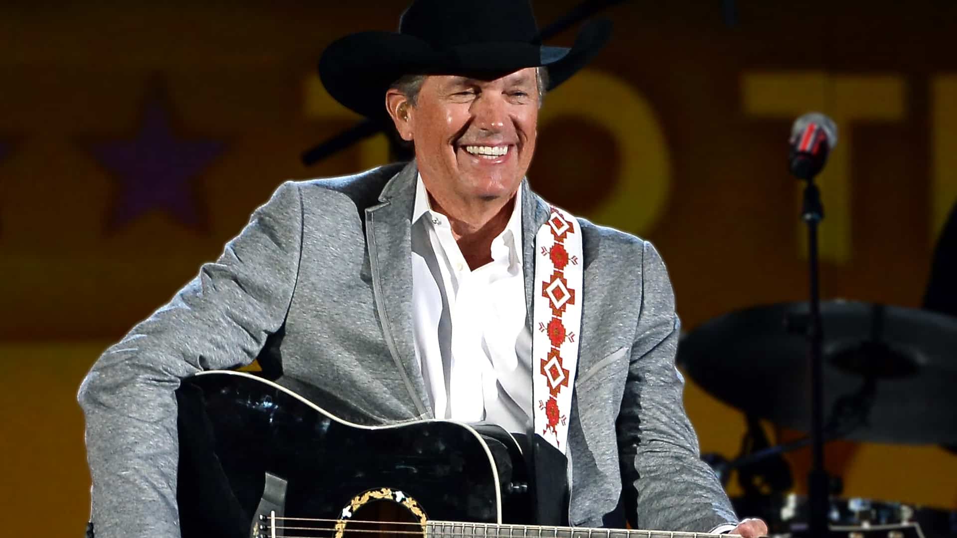 George Strait smiling onstage with his black guitar and cowboy hat, teasing his 2026 return to Lubbock’s Jones AT&T Stadium.