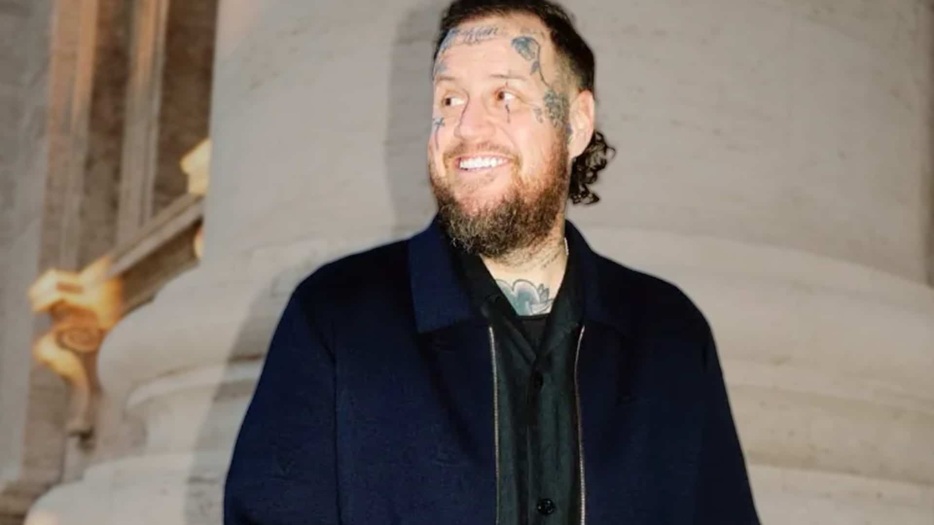 Jelly Roll smiling after massive 200-pound weight loss, showing off a trimmed beard and sharper jawline while joking about needing cosmetic surgery or a facelift in his bold new transformation.