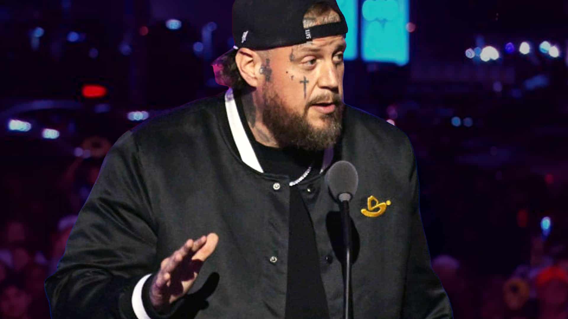 Jelly Roll stands at a microphone wearing a black jacket and backward cap during an event, captured in a powerful image that mirrors the frustration he later described after being treated like a criminal at a Louis Vuitton store in Sydney.