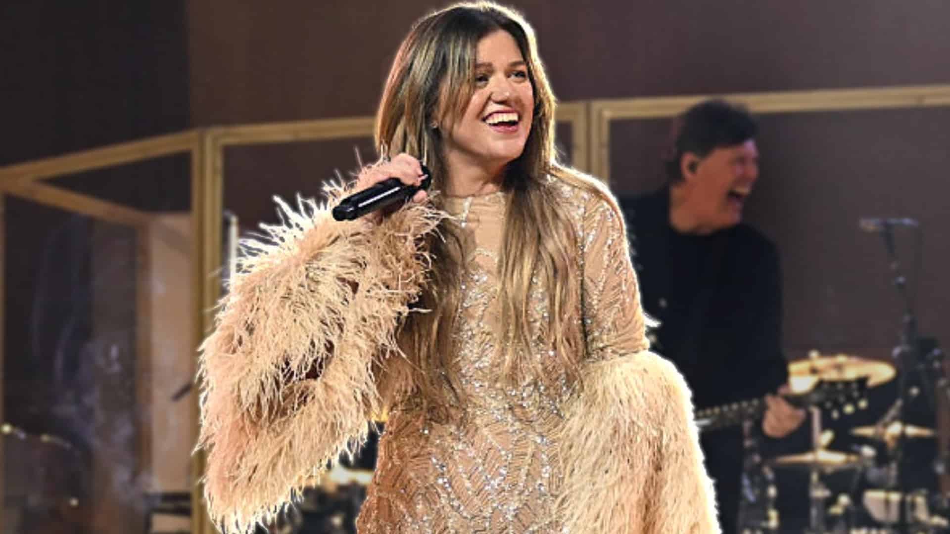 Kelly Clarkson laughing onstage in a sparkling feathered outfit after recalling how a former manager told her to get a boob job.