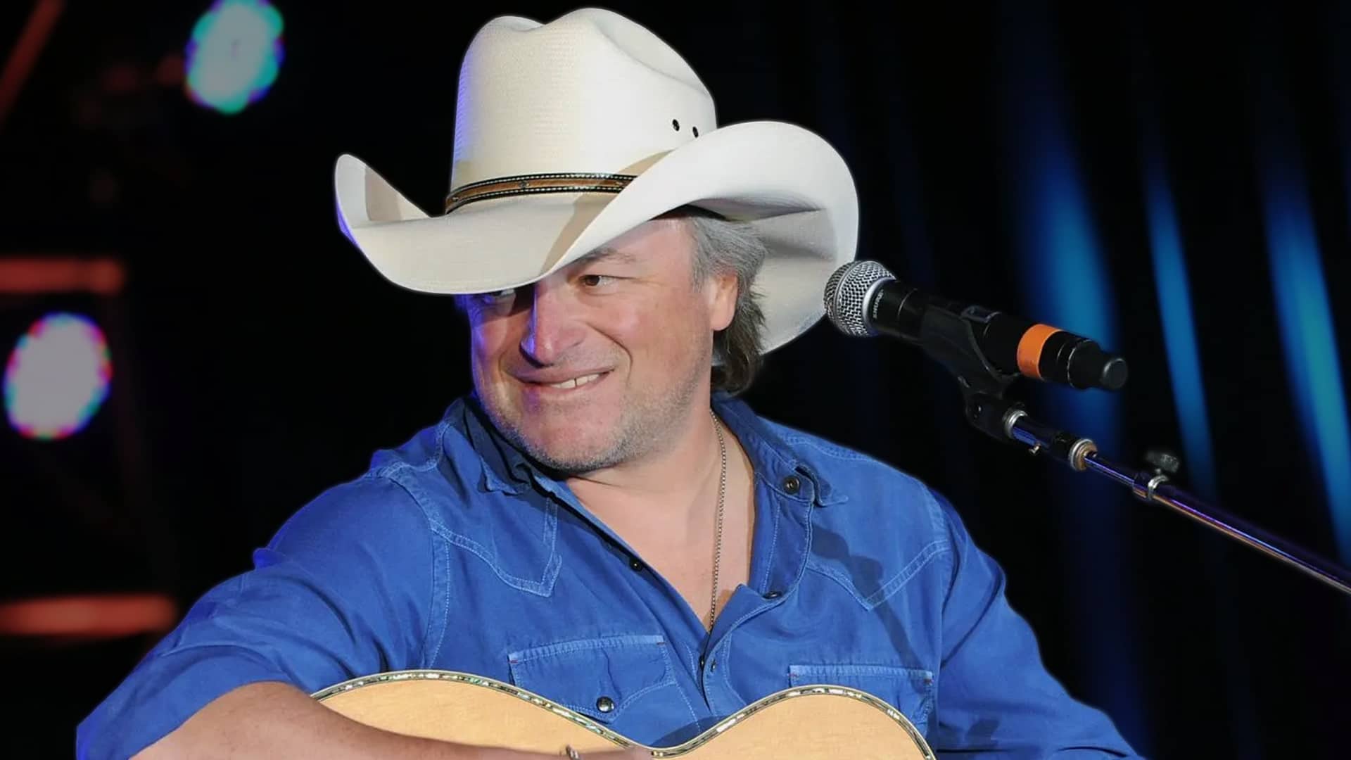 Mark Chesnutt on stage with a guitar and microphone, wearing a white cowboy hat and blue shirt, following news that he was hospitalized and had to postpone tour dates.