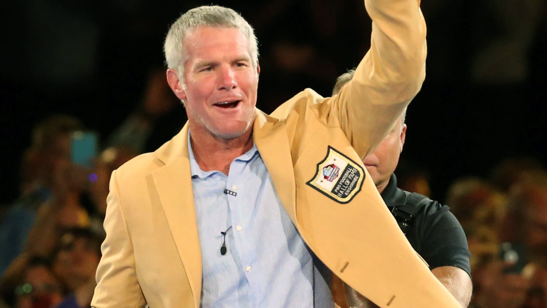 Retired NFL quarterback Brett Favre, wearing a gold Hall of Fame jacket, raises his arm to the crowd, reflecting the authority and pride behind his comments about wanting a patriotic country artist to perform at the Super Bowl.