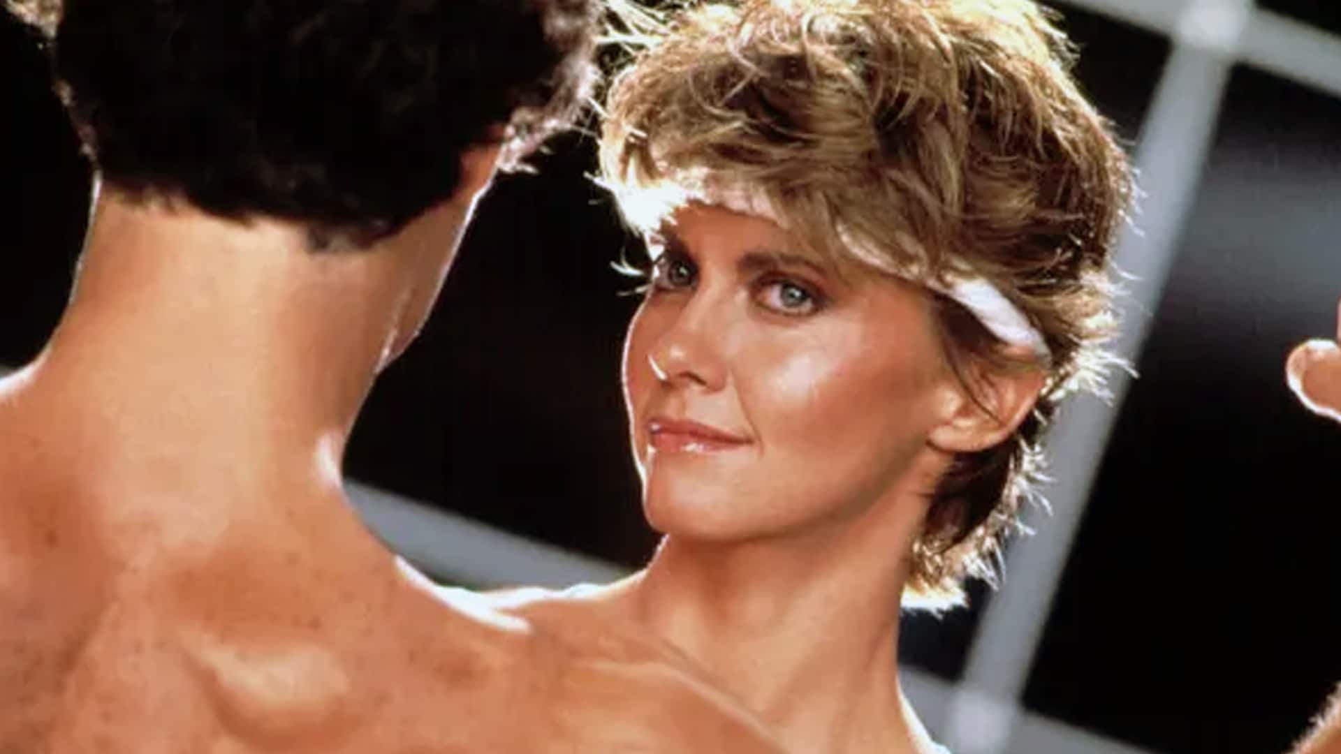Olivia Newton-John with blonde 80s hair and headband looking up at muscular man in black during steamy Physical music video scene from 1981 global No 1 hit she almost scrapped.