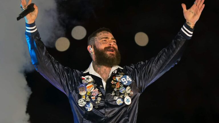 Post Malone raising both arms in triumph during his 2025 Cowboys vs Chiefs Thanksgiving halftime show at AT&T Stadium, honoring Marshawn Kneeland with a No. 94 pin on his custom Dallas jacket.