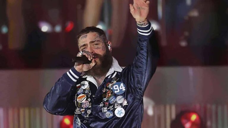 Post Malone performing at the 2025 Cowboys vs Chiefs Thanksgiving halftime show at AT&T Stadium, raising one arm in a pin-covered Cowboys jacket during his high-energy F-1 Trillion set.