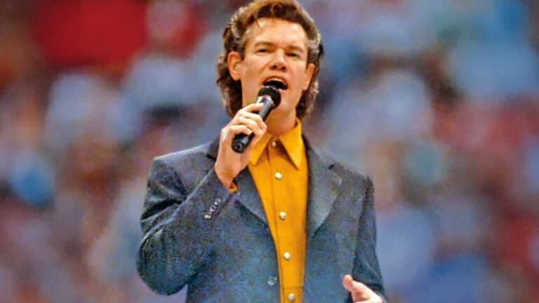 Randy Travis performing at the 1998 Dallas Cowboys Thanksgiving halftime show, rising dramatically from the stage in a blue jacket and yellow shirt, singing into a microphone with the stadium crowd in the background.