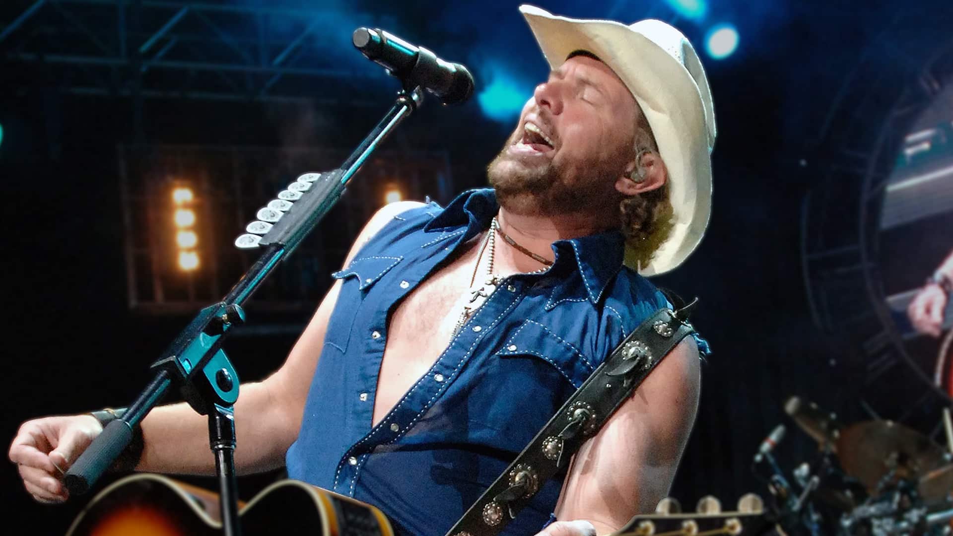 Toby Keith passionately performing "I Wanna Talk About Me" live on stage in a white cowboy hat and sleeveless denim shirt – the 2001 No. 1 hit originally written for Blake Shelton.