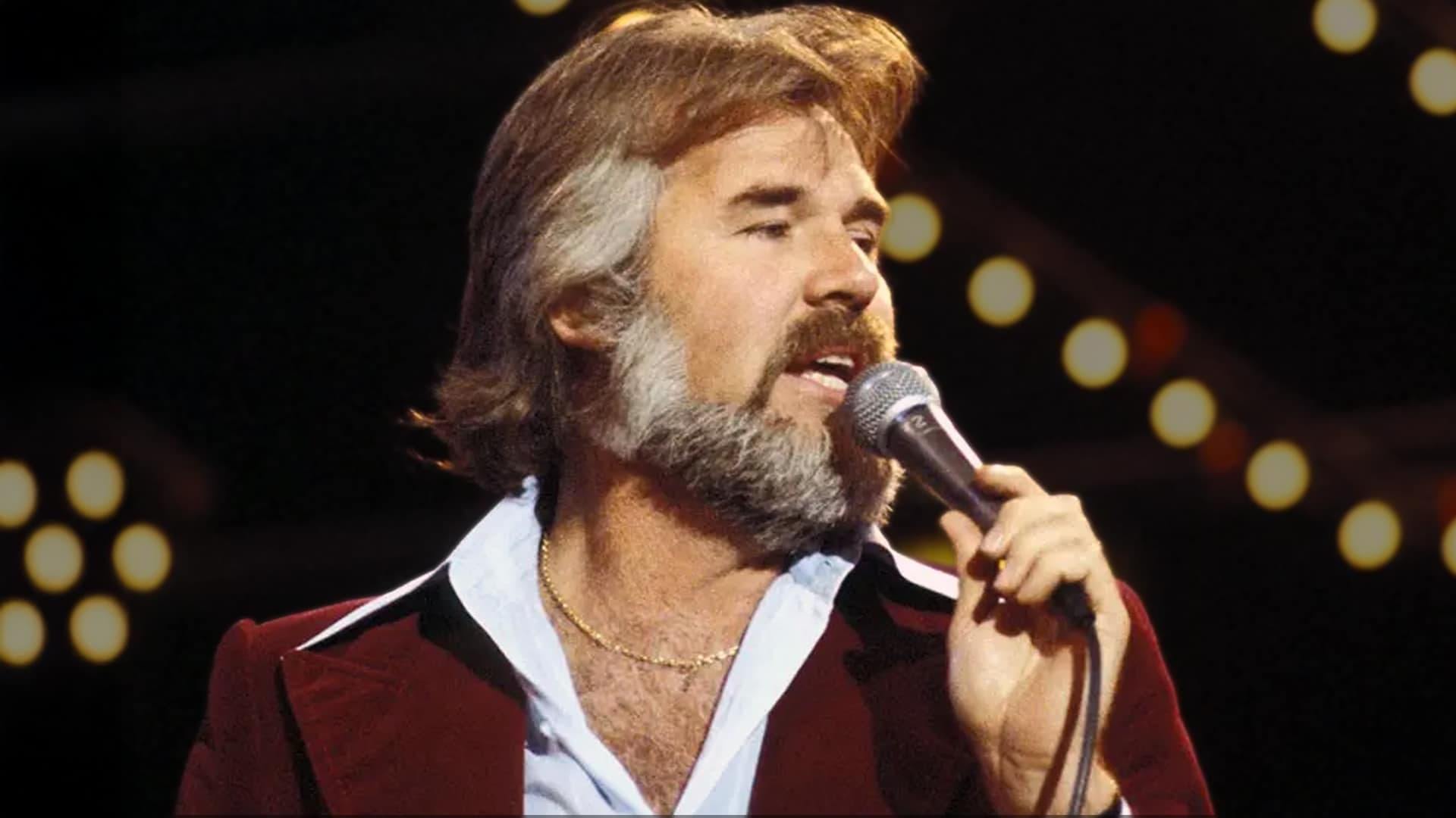 Kenny Rogers singing under warm stage lights, capturing the swagger tied to the legendary backstory behind the hit that became his signature.