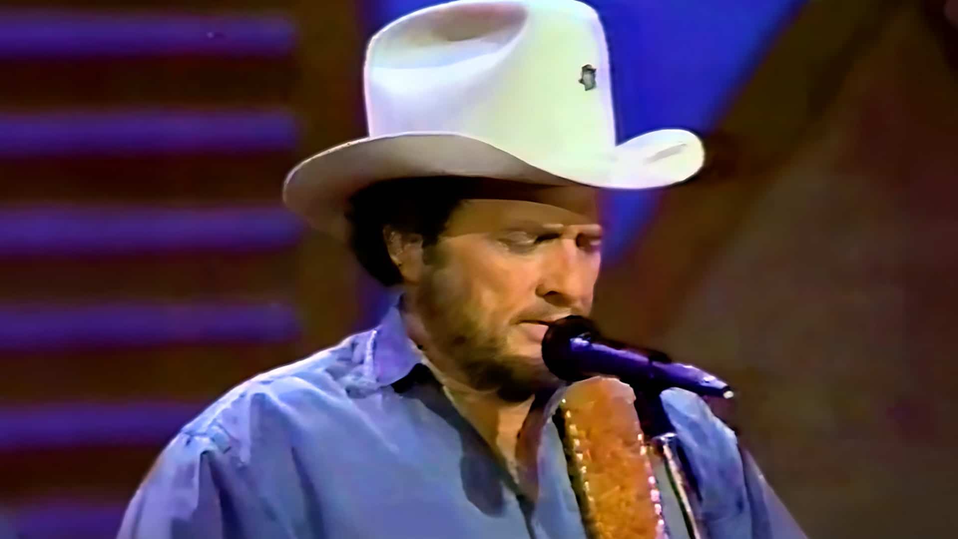 Merle Haggard in signature white cowboy hat and light blue shirt singing into microphone holding guitar during live performance of Twinkle Twinkle Lucky Star his 38th and final Billboard country No 1 from 1987 Chill Factor album.