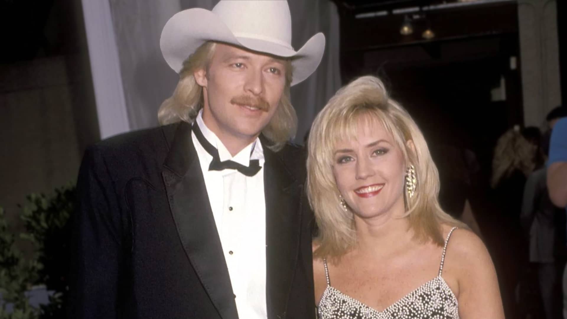Alan Jackson and wife Denise Jackson in a classic 1990s photo, Alan in white cowboy hat and tuxedo, Denise in sparkling dress, celebrating their enduring marriage that began with Alan's wild stunt of hiding in her car backseat at Dairy Queen to get her attention.