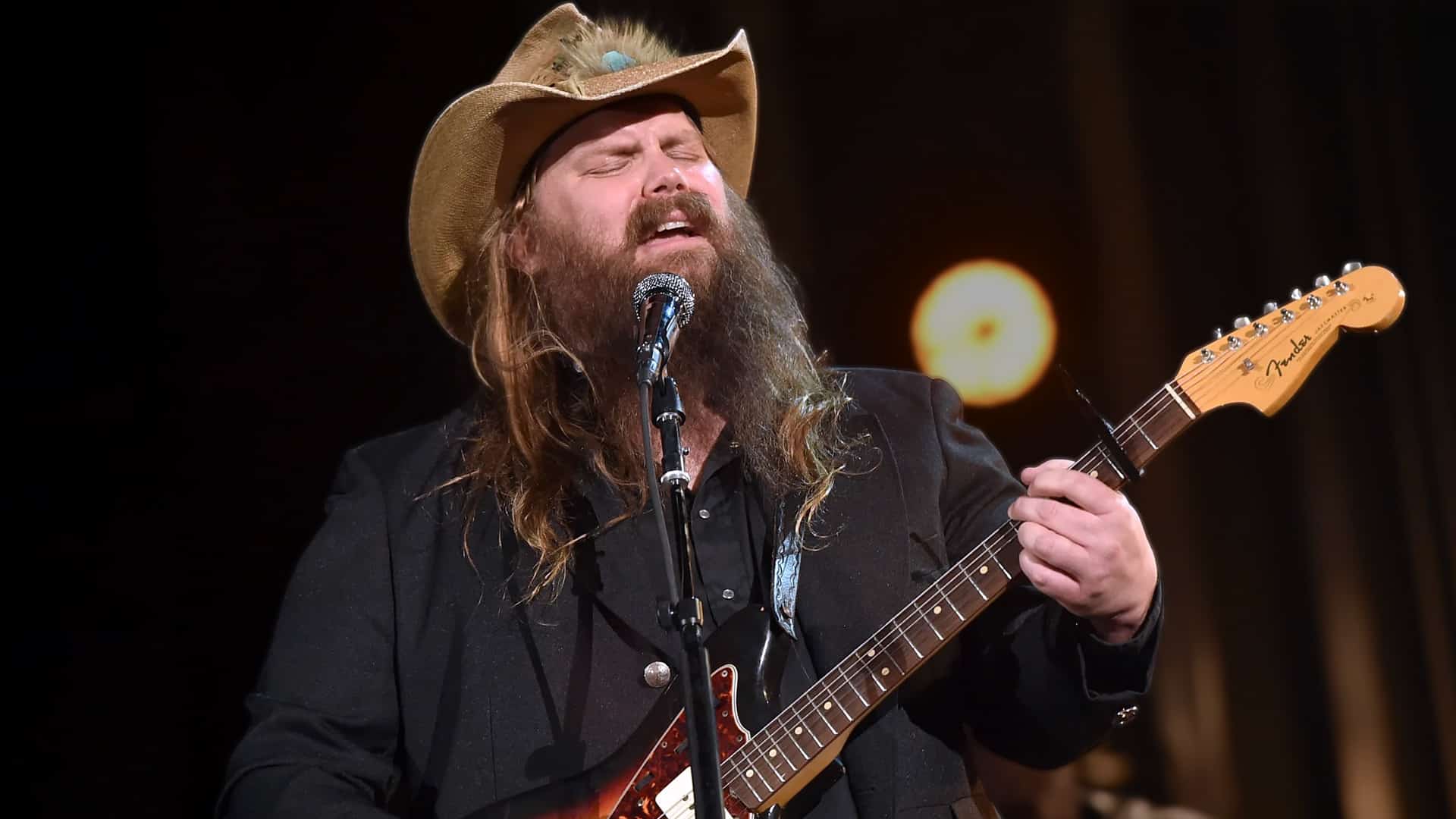Chris Stapleton performing his iconic "Tennessee Whiskey" live on stage in a cowboy hat and black suit, passionately singing and playing a sunburst Fender guitar as the 2015 hit celebrates 10 years and still outcharts newer country releases on Apple Music's 2025 Top 100.