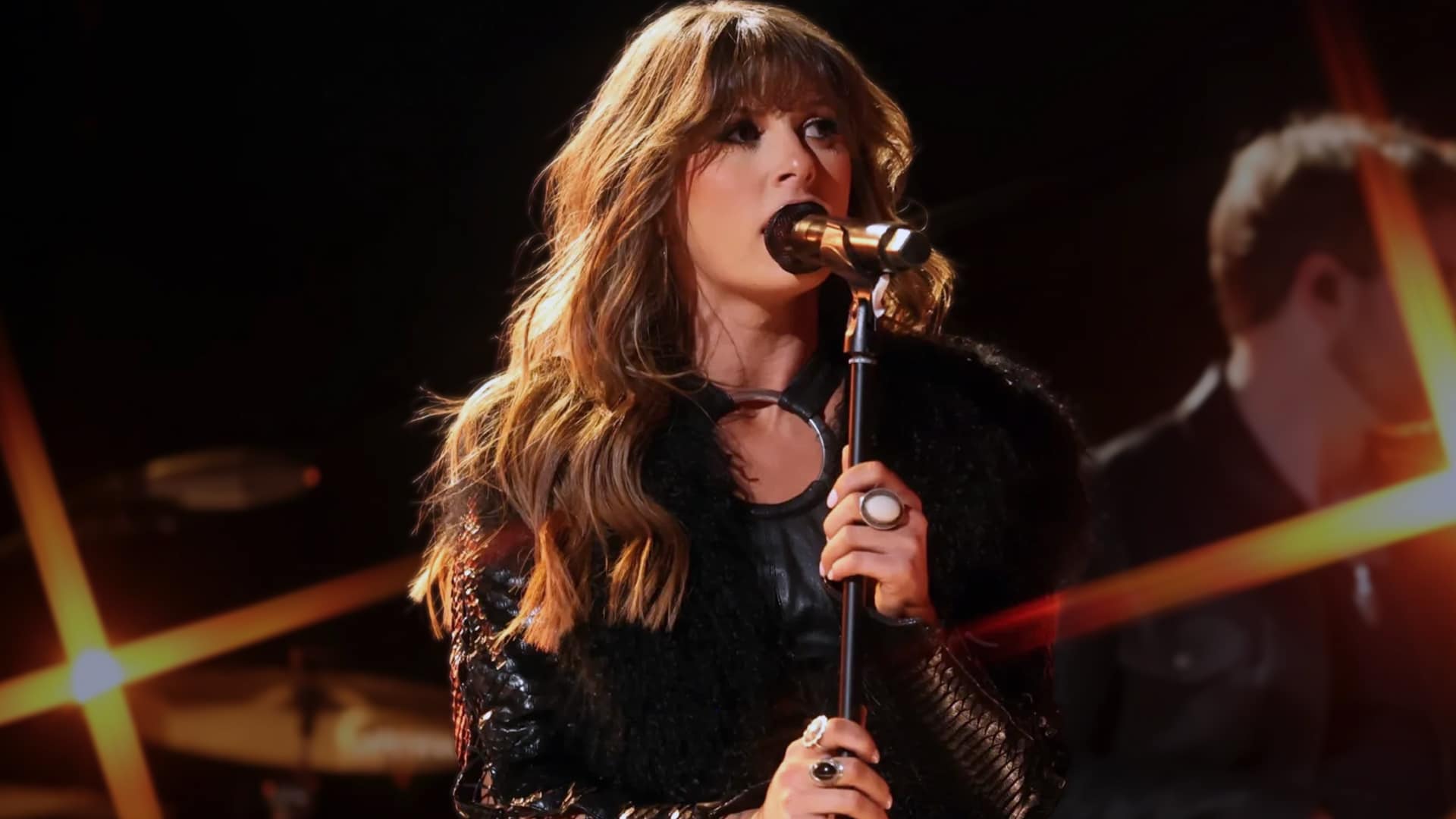 Ella Langley performs energetically on stage in black fuzzy jacket and leather accents, holding a microphone, as her single "Choosin' Texas" becomes the fastest solo female song to crack Country Radio's Top 10 in a decade in just eight weeks during 2025.