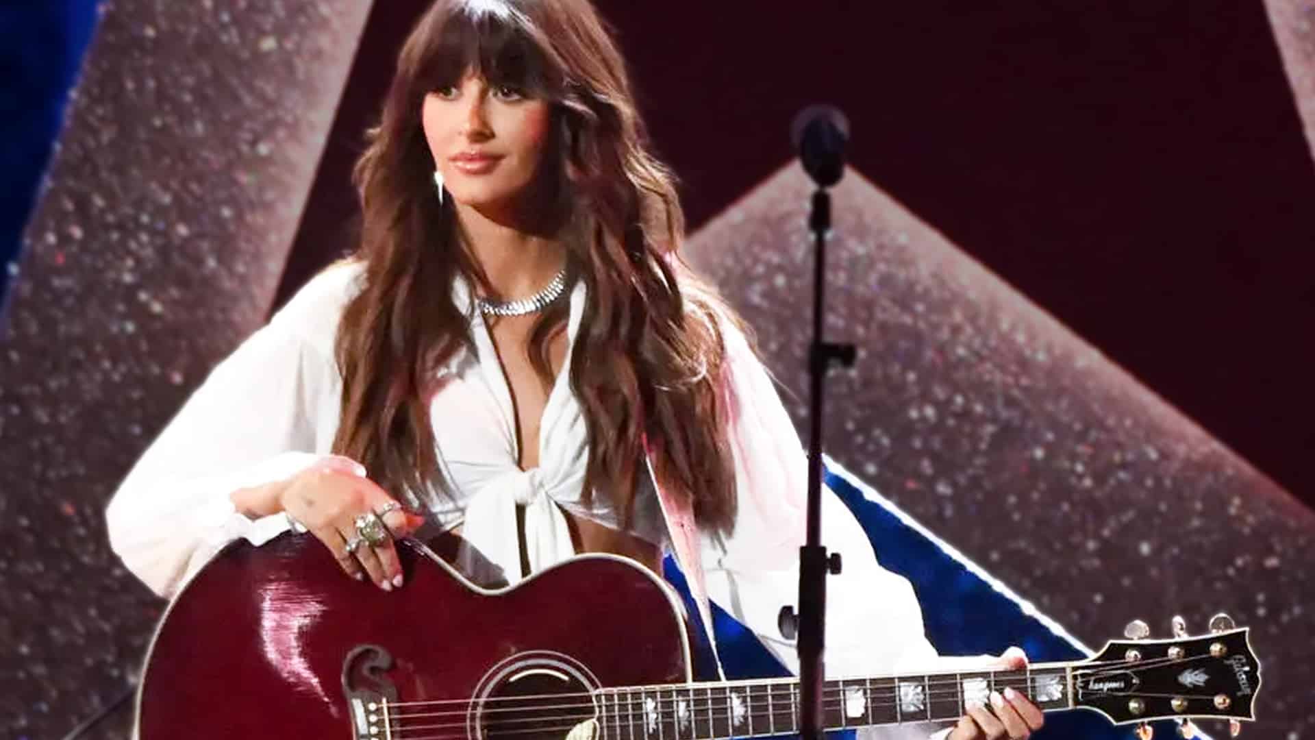 Ella Langley celebrates her first career No. 1 hit "Choosin' Texas" on Billboard Hot Country Songs, performing on stage with a red acoustic guitar, white blouse, and sparkling choker while knocking Morgan Wallen from the top spot in late 2025.