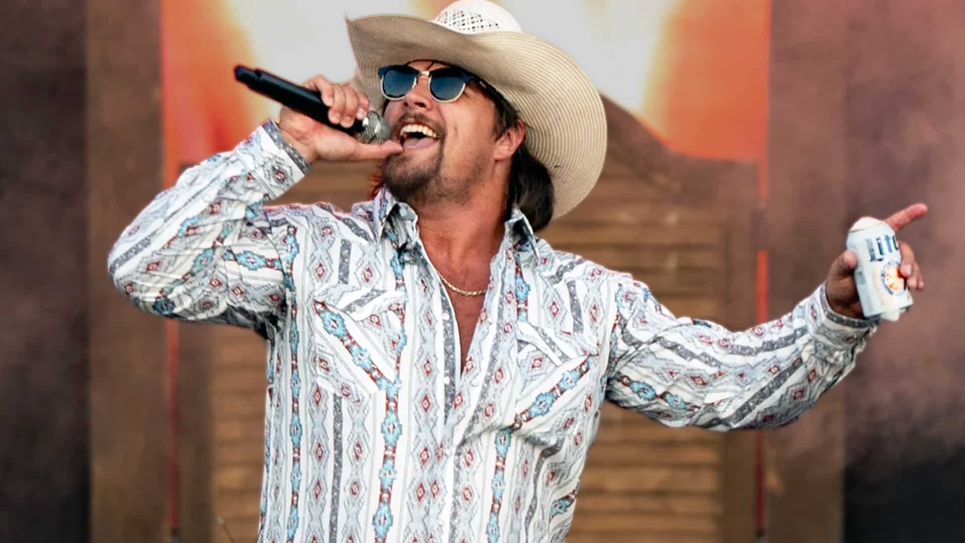 Gavin Adcock performing live on stage in a white cowboy hat, sunglasses, and patterned western shirt, holding a mic in one hand and a Miller Lite in the other, revealing that Alan Jackson's "Remember When" is the one country song that still brings him to tears every time.