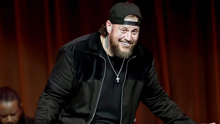Jelly Roll smiles on stage in black cap and velvet jacket with cross necklace and tattoos, revealing in 2025 how losing over 200 pounds unexpectedly cured his lifelong color blindness, allowing him to see vibrant colors like purple for the first time.