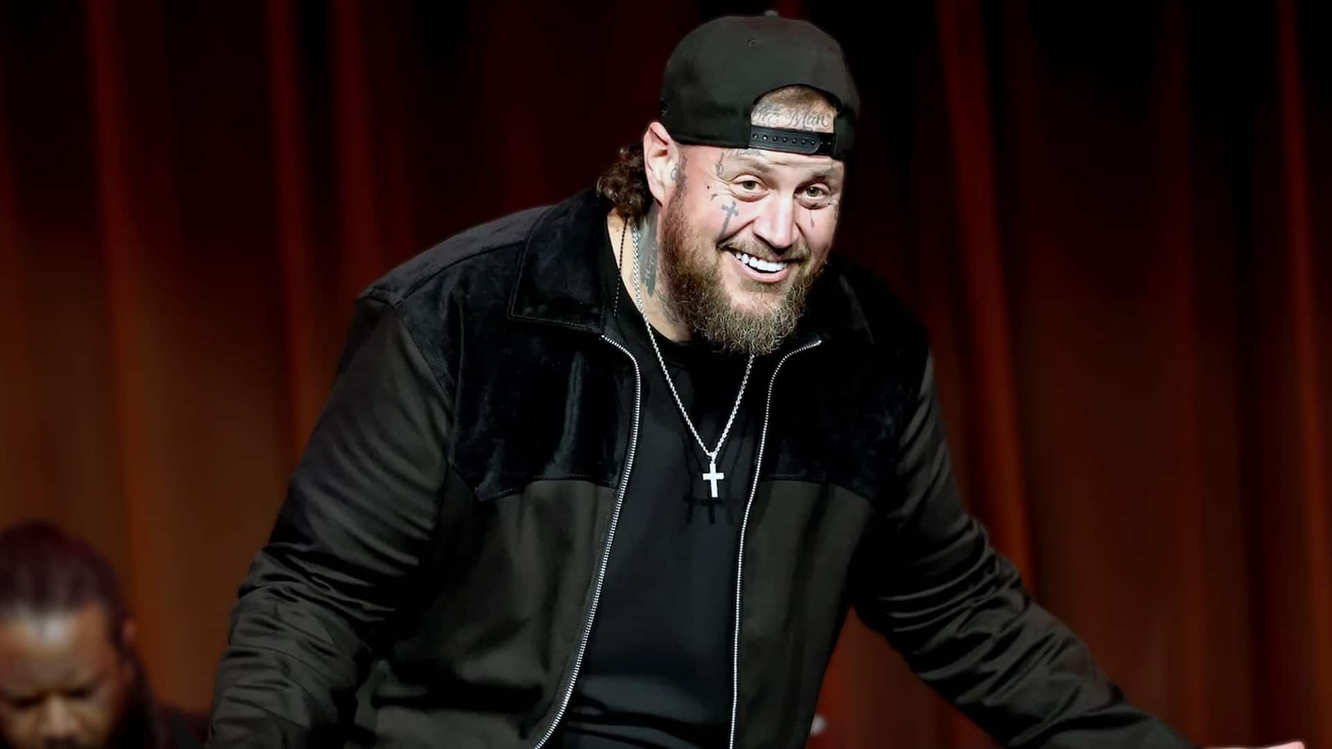 Jelly Roll smiles on stage in black cap and velvet jacket with cross necklace and tattoos, revealing in 2025 how losing over 200 pounds unexpectedly cured his lifelong color blindness, allowing him to see vibrant colors like purple for the first time.