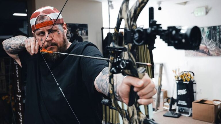 Jelly Roll aiming a compound bow indoors in a patriotic cap and black tee with visible tattoos, advocating for restored firearm rights to hunt as a reformed felon, sharing his redemption story and push for second chances in 2025.