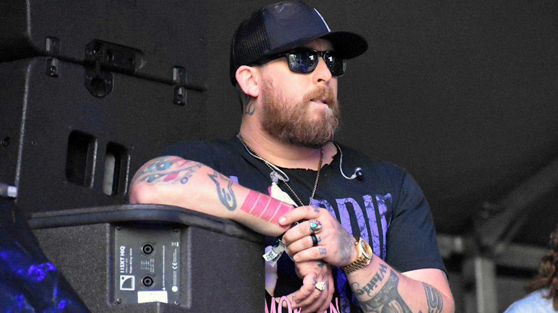 Jesse Keith Whitley, son of Keith Whitley and Lorrie Morgan, leans on stage equipment in black cap and sunglasses with tattoos visible, revealing his 2025 hospitalization for severe pancreatitis and life-changing decision to embrace sobriety and faith.